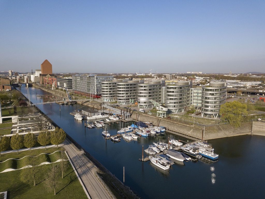 Drohnenaufnahme eines kleinen Hafens mit modernen Gebäuden und umliegender Umgebung im Hintergrund - professionelle Architekturfotografie