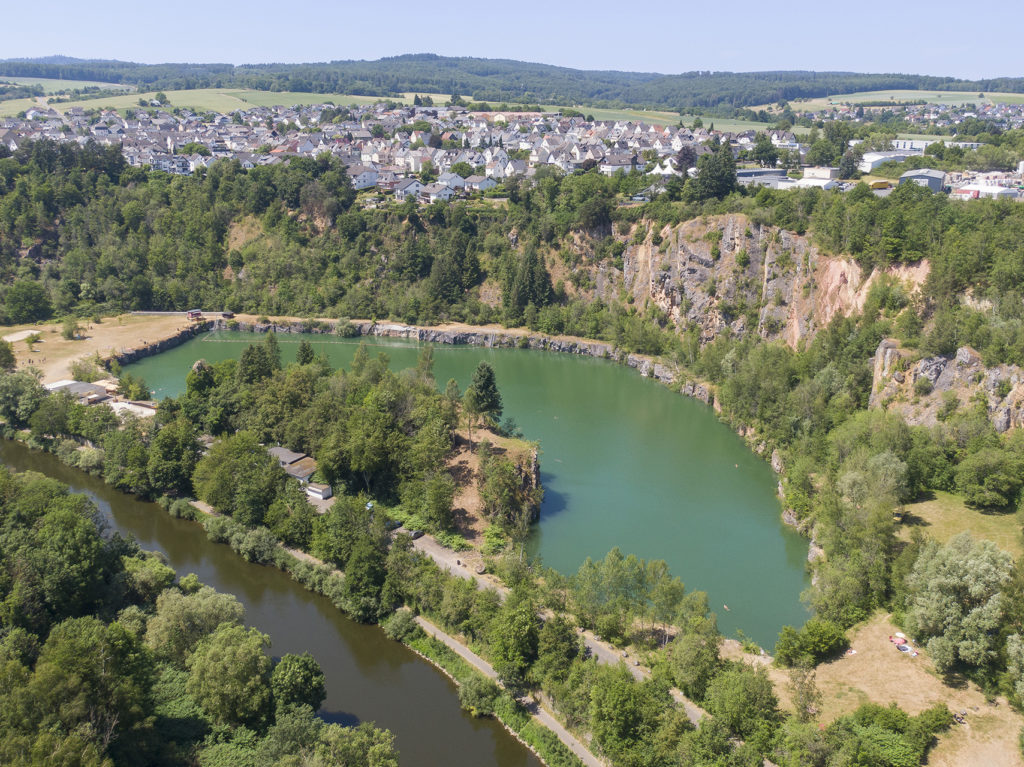 Drohnenaufnahme einer sonnigen Landschaft mit Klippe, Baggersee in Diez und der Lahn - professionelle Tourismusfotografie